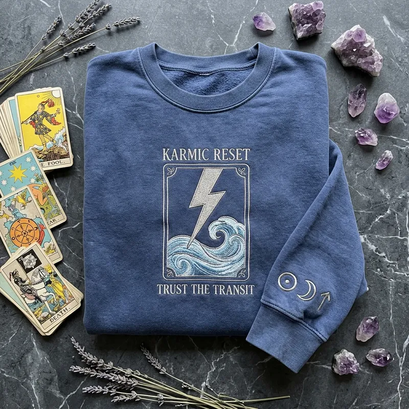 April 7 2026 Astrology: The Karmic Reset & Cozy Custom Apparel Close up macro shot showing the fine-line metallic silver embroidery thread of a lightning bolt striking a swirling wave on the Karmic Reset sweatshirt