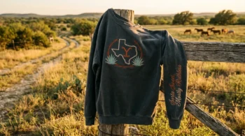 Dutton Ranch season 1 details on a vintage washed charcoal embroidered sweatshirt draped on a rustic fence.
